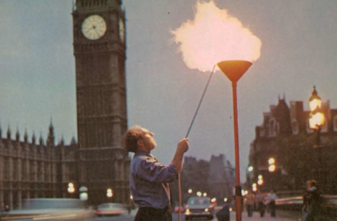 A man lighting a tall gas flame beside Big Ben in London.