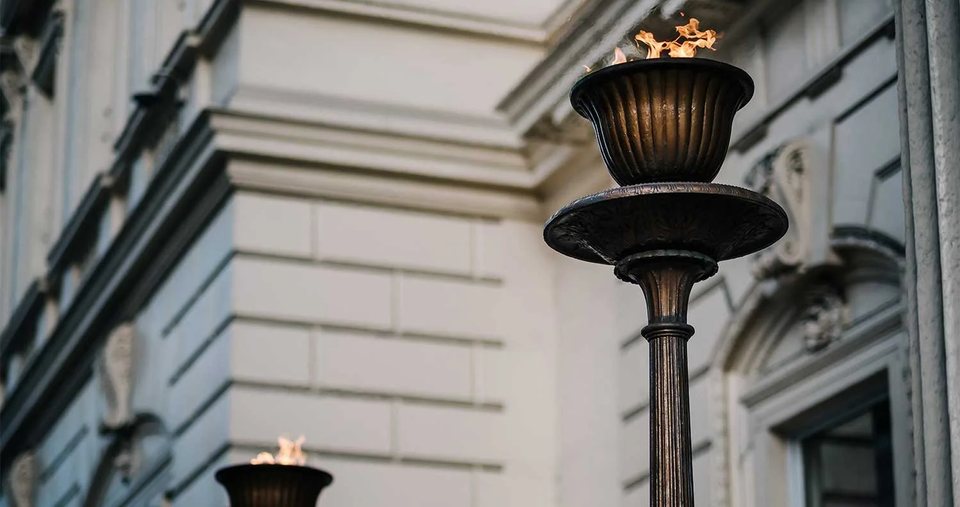 A gas flame burning from a lamp on a stone pillar outside a grand building.